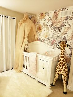 a giraffe standing next to a baby crib in a room with floral wallpaper