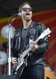 a man in black jacket and sunglasses playing an electric guitar on stage at a music festival