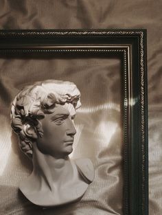 a white busturine in a black frame on a gray cloth covered background with the shadow of a man's head