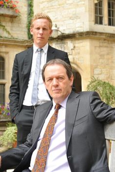 two men in suits and ties sitting on a bench next to each other, one is looking at the camera