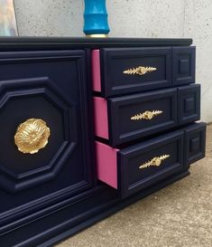 a blue and pink dresser with gold accents on it's drawers, next to a vase