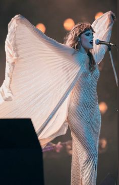 a woman in white dress standing on stage with her arms spread out to the side