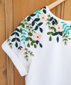 a white shirt with flowers and leaves embroidered on the front, hanging from a wooden hanger