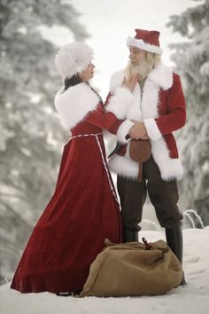 two people dressed as santa and mrs claus standing in the snow with a bag on their lap