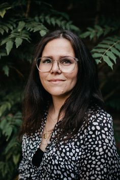 a woman with glasses standing in front of some trees and plants, smiling at the camera