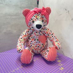 a teddy bear with a pink bow sitting on a purple tableclothed surface next to a gray wall