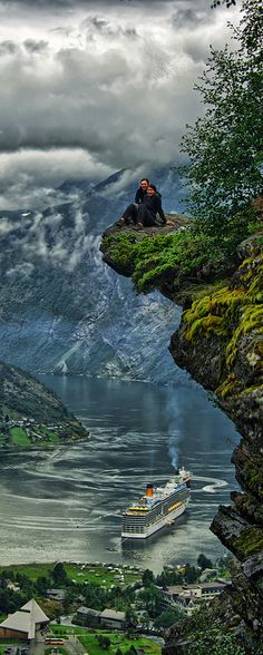 two people sitting on the edge of a cliff looking at a boat in the water