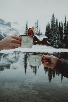 two people holding coffee cups in front of a mountain lake with trees and snow on the ground