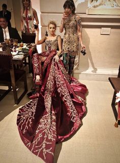 two women dressed in red and gold gowns standing next to each other at a dinner table