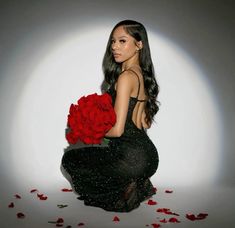 a woman in a black dress holding a red rose with petals scattered around her and sitting on the floor