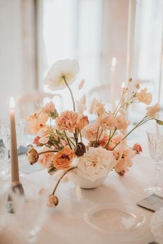 an arrangement of flowers in a white vase on a table with silverware and candles