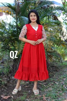 a woman wearing a red dress standing in front of some plants and trees with her hands on her hips