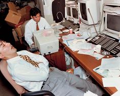 two men sitting at a desk in front of computer monitors and other office equipment with boxes on the floor