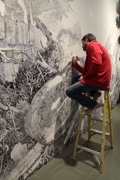 a man sitting on a stool in front of a wall painting with black and white images