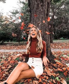 a woman sitting on the ground next to a tree with leaves all around her and laughing