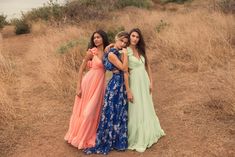 three women in long dresses standing on the side of a dirt road next to dry grass