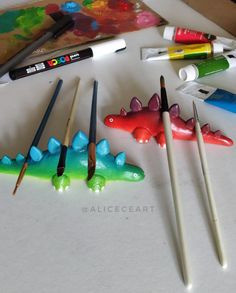 A photo of brightly coloured dinosaurs. They are made so that paintbrushes can be rested in the gaps between the spikes along the back. Around the dinosaurs are some paint tubes and a Posca paint pen Air Dry Clay Unique Ideas, Clay Worm Paint Brush Holder, Worm Paintbrush Holder, Clay Paint Holder, Air Dry Clay Paint Pallet, Paint Brush Rest Diy, Clay Paintbrush Rest, Polymer Clay Paint Brush Holder, Air Dry Clay Paintbrush Holder