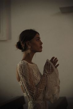 a woman standing in front of a window wearing a white dress and holding her hands together