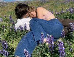 two people are laying in the middle of a field with purple flowers on it and one person is hugging her head