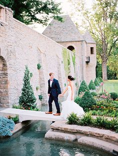 a bride and groom walking across a bridge in front of an old brick building with water running through it