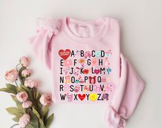 a pink sweater with alphabets and hearts on it next to some flowers, including roses