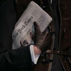 a man in black jacket and brown gloves reading a newspaper with his hand on top of the newspaper