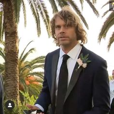 a man in a suit and tie standing next to a palm tree with other people behind him