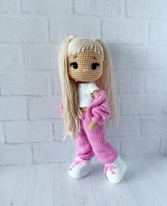 a crocheted doll with long blonde hair wearing pink pants and white shirt standing in front of a brick wall