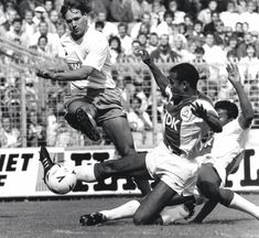 two men are playing soccer in front of an audience at a sporting event, one is diving for the ball while the other tries to block it