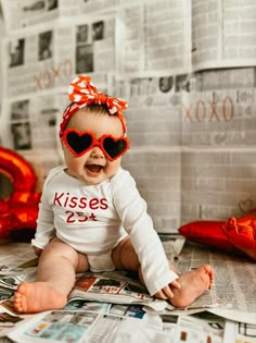 a baby sitting on the floor wearing red sunglasses and a bow in her hair, with newspaper behind her