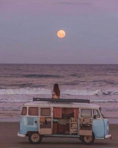 a woman sitting on top of an old vw bus next to the ocean at sunset