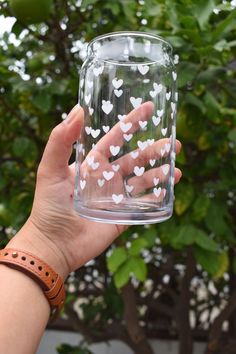 a person holding up a glass with white hearts on it in front of a tree