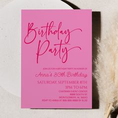 a pink birthday party card sitting on top of a white table next to a feather