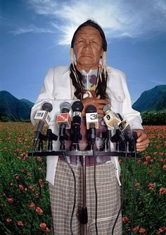 an old man standing in front of a bunch of microphones with mountains in the background