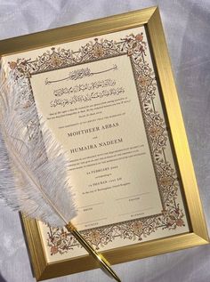 a white feather laying on top of a paper with an ornate border and gold frame