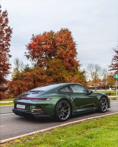 a green sports car driving down the road in front of some trees with orange leaves