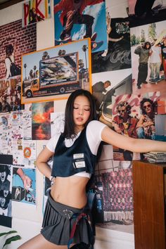 a young woman posing in front of a wall covered with pictures and posters, leaning on a desk