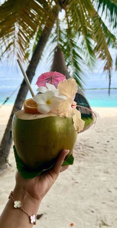 a person holding up a coconut drink with flowers in it on a beach near the ocean