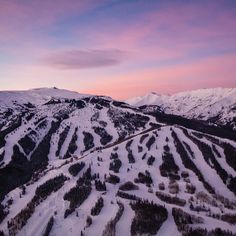 the mountains are covered in snow and trees under a pink sky with clouds over them