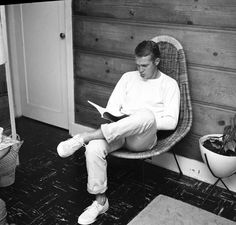 a man sitting on a chair reading a book in front of a wooden wall and potted plant