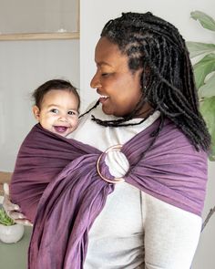 a woman holding a baby wrapped in a purple wrap and smiling at the camera while standing next to a potted plant