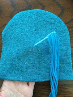 a hand holding a blue knitted beanie on top of a wooden table next to a pair of scissors