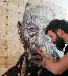 a man in black shirt writing on wall next to large drawing of an old man