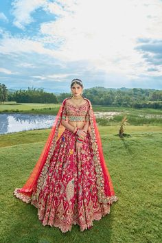 a woman in a red and gold bridal gown standing on the grass near a lake