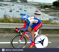 a man riding his bike down the road with boats in the water and buildings behind him