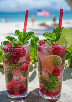 two glasses filled with ice and raspberries next to each other on the beach