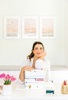 a woman sitting at a desk with stacks of books in front of her and flowers on the table