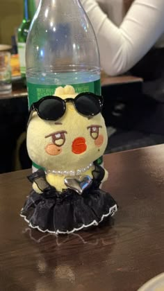 a stuffed animal wearing sunglasses and a dress sitting on top of a table next to a bottle