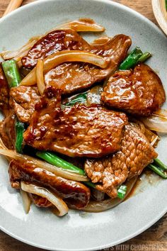 a white plate topped with meat and onions on top of a wooden table next to chopsticks