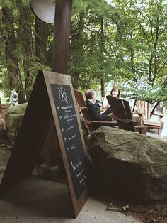 two people are sitting at a table in the woods and one person is writing on a blackboard